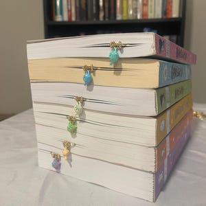 Puede incluir: Una pila de seis libros con marcapáginas de metal sujetos a los bordes. Cada marcapáginas presenta un pequeño dije de tetera de colores en tonos azul claro, azul brillante, blanco, verde, amarillo y morado, con cuentas de tono dorado.
