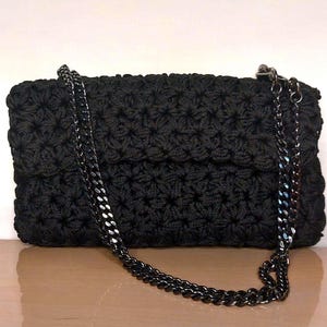 Könnte beinhalten: Eine schwarze Häkeltasche mit Kettenriemen. Die Tasche hat ein strukturiertes, sternförmiges Muster auf der Oberfläche. Der Kettenriemen ist dunkelgrau, was einen Kontrast zum einfarbigen Schwarz der Tasche bildet.