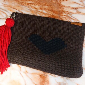 Peut inclure: Une pochette rectangulaire marron avec un motif en forme de cœur noir. La pochette est ornée d'un pompon rouge attaché à une tirette de fermeture éclair. Le sac est en tricot.