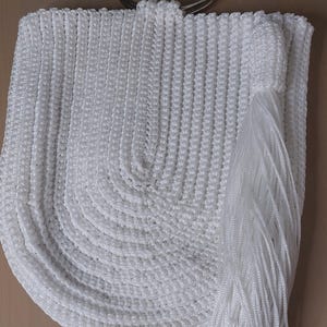 May include: White crocheted handbag with a silver metal handle. The bag features a rounded bottom and a long tassel. The crochet pattern is visible, and the bag appears to be made of a soft, textured material.