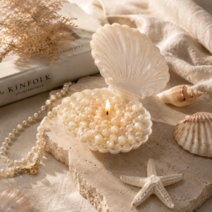 May include: A white shell-shaped candle holder containing numerous small, round, cream-colored pearls and a single lit wick in the center. The open shell is positioned alongside a long strand of matching pearls and a small white starfish.