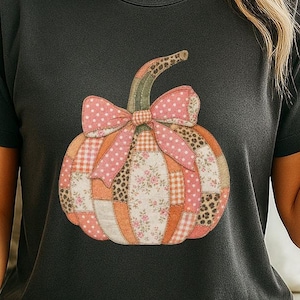May include: A dark gray short-sleeve t-shirt featuring a central graphic of a pumpkin made of various patchwork patterns including pink polka dots, leopard print, and floral designs, topped with a large pink polka dot bow tied at the stem.