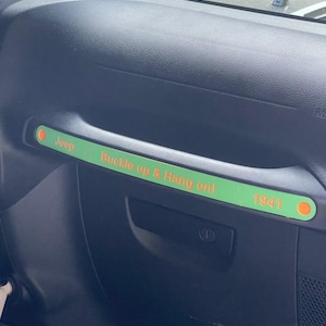 May include: A black interior car handle with a green accent. The green accent has the text "Jeep Buckle up & Hang on! 1941" in orange lettering. The handle is part of a vehicle's dashboard.