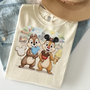 May include: Cream-colored t-shirt featuring Chip and Dale with treats in front of a Disney castle. Chip holds a blue ice cream, and Dale holds a Mickey Mouse ice cream and a soft-serve cone. A Mickey Mouse-shaped cookie is also visible.