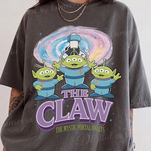 May include: A charcoal gray short-sleeve t-shirt featuring a graphic of three green three-eyed aliens in blue space suits under a swirling purple and pink galaxy. The text reads THE CLAW and THE MYSTIC PORTAL AWAITS in purple and green lettering.