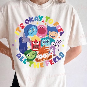 May include: Cream-colored t-shirt featuring a colorful graphic with the text "IT'S OKAY TO FEEL ALL THE FEELS" and cartoon characters representing different emotions. The design includes characters in various colors, such as blue, red, green, and orange.