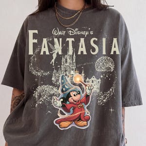 May include: A charcoal gray short-sleeve t-shirt featuring a graphic print of Mickey Mouse as a sorcerer from Walt Disney's Fantasia. The design includes a castle, Tinker Bell, and the text Walt Disney's Fantasia in a large, cream-colored font.