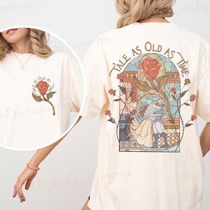 May include: Cream-colored t-shirt featuring a rose and the text "Tale as Old as Time." The back of the shirt displays a stained-glass design with the same phrase and a romantic scene. The shirt has short sleeves and a relaxed fit.