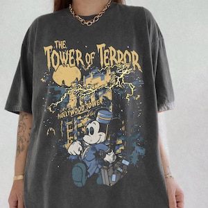 May include: Dark gray t-shirt featuring a graphic of Mickey Mouse in a bellhop uniform running in front of the Hollywood Tower Hotel. The text "The Tower of Terror" is above the image, with lightning bolts and a yellow moon in the background.