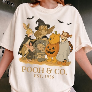 May include: A cream-colored short-sleeve t-shirt featuring a graphic of cartoon characters dressed in Halloween costumes, including a pumpkin, a mummy, and a witch. The text "POOH & CO. EST. 1926" is printed in brown below the characters.