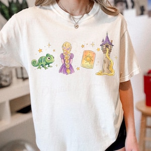 Pode incluir: Uma t-shirt de manga curta em tom branco sujo com uma impressão gráfica de um camaleão verde, uma personagem com um longo penteado trançado loiro num vestido roxo, uma lanterna amarela brilhante e uma torre de pedra alta com uma trança dourada.