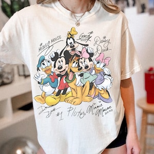 Puede incluir: Una camiseta de manga corta color crema con un estampado gráfico de Mickey Mouse, el Pato Donald, Goofy, Pluto, Minnie Mouse y Daisy Duck. El diseño incluye etiquetas de texto estilo manuscrito para cada personaje y el texto #1.