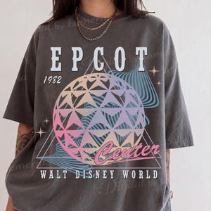 May include: A charcoal gray short-sleeve t-shirt featuring a graphic of the Epcot geodesic sphere in pastel pink, purple, and blue. The text reads EPCOT 1982 Center WALT DISNEY WORLD, accompanied by star icons and abstract blue wave patterns.