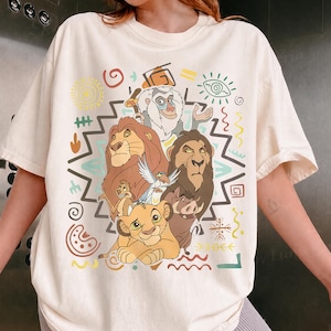 Op de afbeelding: Een crèmekleurig T-shirt met korte mouwen en een kleurrijke grafische print van diverse geanimeerde leeuwen- en dierfiguren. Het ontwerp bevat een centrale groep personages omringd door geometrische vormen, krullen en tribale patronen.