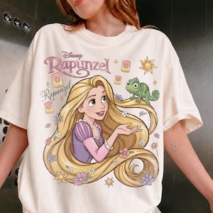 May include: A cream-colored t-shirt featuring a graphic print of the character Rapunzel with long blonde hair, a purple dress, and a green chameleon. The design includes the text Disney Rapunzel, floating lanterns, a sun icon, and small flowers.
