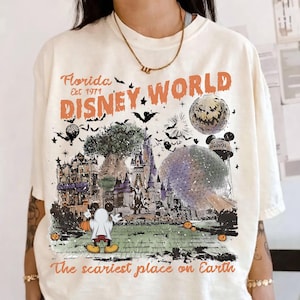 May include: An off-white t-shirt featuring a graphic print with the text Florida Est 1971 DISNEY WORLD The scariest place on Earth. The design includes a castle, a large sphere, a ghost-costumed mouse, bats, and a spooky moon illustration.