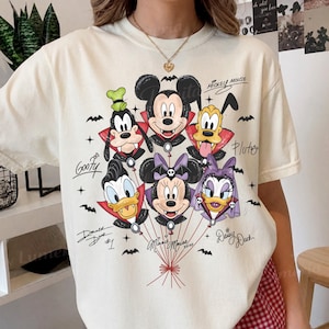 Puede incluir: Una camiseta de manga corta color crema con un gráfico de seis personajes de Disney vestidos de vampiros, incluyendo a Mickey Mouse, Minnie Mouse, Donald Duck, Daisy Duck, Goofy y Pluto, todos unidos a cuerdas de globos con murciélagos.