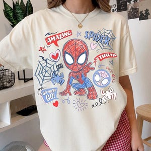 Peut inclure: Un t-shirt à manches courtes de couleur crème arborant un motif de Spider-Man rouge et bleu dans un style esquissé. Le design comprend les textes AMAZING, SPIDER, THWIP, WEB-HEAD et GO-WEBS-GO, ainsi que des toiles d'araignée et des cœurs.