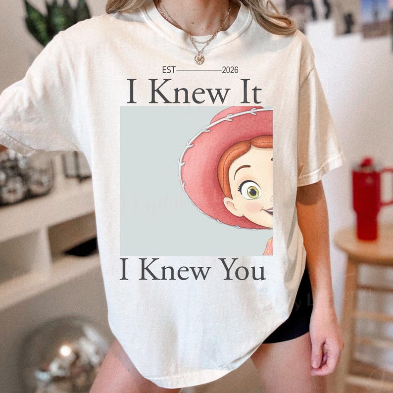 Pode incluir: Uma t-shirt de manga curta cor de creme com um gr&aacute;fico de uma personagem de desenho animado a usar um chap&eacute;u de cowgirl cor-de-rosa. O texto "EST 2026", "I Knew It" e "I Knew You" est&aacute; impresso a preto acima e abaixo da imagem.