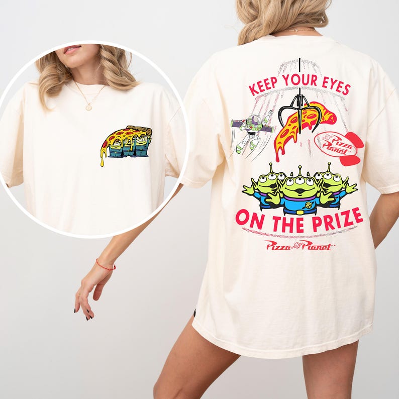 May include: Cream-colored t-shirt featuring a Pizza Planet design. The front has a pizza slice graphic with aliens. The back displays the text "KEEP YOUR EYES ON THE PRIZE" with Buzz Lightyear, a claw machine, and the Pizza Planet logo.