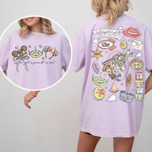 May include: A lilac t-shirt with cartoon character illustrations on the front and back. The front shows a candy, alien, and bow design with the text "You've got a friend in me!". The back has a hat, Buzz Lightyear, Woody, and other character graphics.