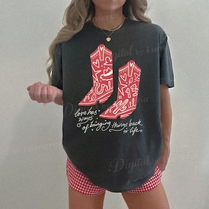 Puede incluir: Una camiseta de manga corta gris carb&oacute;n con un gr&aacute;fico de dos botas vaqueras rojas decoradas con patrones de l&iacute;neas blancas de estrellas, corazones y herraduras. Debajo de las botas, el texto dice, Love has ways of bringing things back to life.