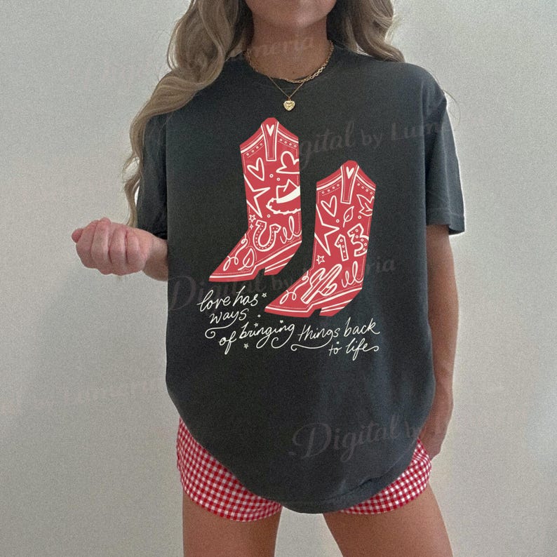 Puede incluir: Una camiseta de manga corta gris carb&oacute;n con un gr&aacute;fico de dos botas vaqueras rojas decoradas con patrones de l&iacute;neas blancas de estrellas, corazones y herraduras. Debajo de las botas, el texto dice, Love has ways of bringing things back to life.