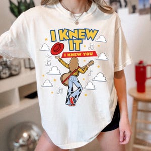 May include: An off-white t-shirt featuring a graphic of a person with long hair playing a guitar, wearing a yellow plaid shirt and cow-print pants. The text reads I KNEW IT and I KNEW YOU, surrounded by red hat, musical notes, stars, and clouds.