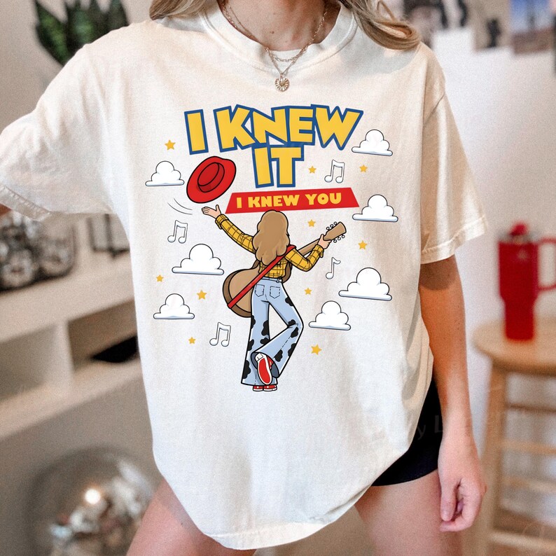 May include: An off-white t-shirt featuring a graphic of a person with long hair playing a guitar, wearing a yellow plaid shirt and cow-print pants. The text reads I KNEW IT and I KNEW YOU, surrounded by red hat, musical notes, stars, and clouds.