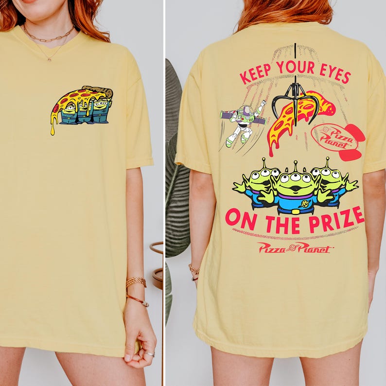 May include: A light yellow t-shirt featuring a cartoon graphic of three green aliens holding a slice of pizza. The back of the shirt has the text "KEEP YOUR EYES ON THE PRIZE" with additional graphics and the "Pizza Planet" logo.