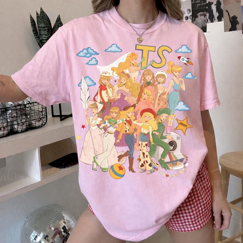 Peut inclure: Un t-shirt rose clair &agrave; manches courtes avec une grande impression graphique repr&eacute;sentant divers personnages de dessins anim&eacute;s dans un style collage. Le motif inclut les lettres TS, une fus&eacute;e, une &eacute;toile de sh&eacute;rif et une balle color&eacute;e.