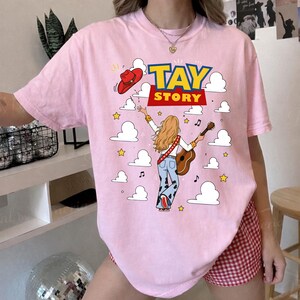 May include: A light pink t-shirt featuring a graphic print of a person with long blonde hair playing an acoustic guitar, wearing cow-print pants and a red cowboy hat. The text "TAY STORY" is printed in blue and yellow above the figure.