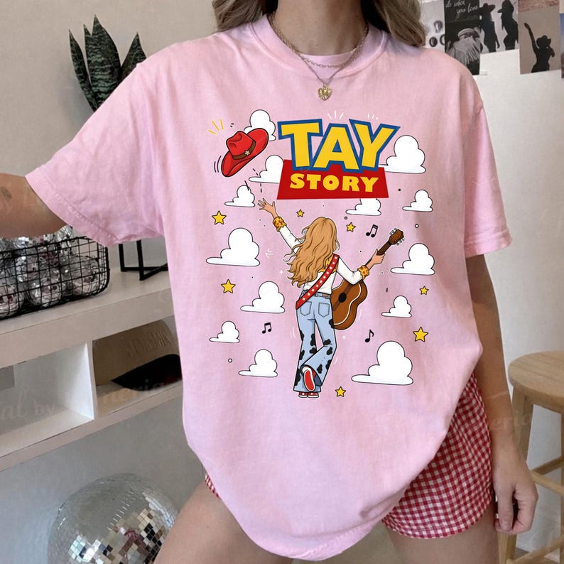May include: A light pink t-shirt featuring a graphic print of a person with long blonde hair playing an acoustic guitar, wearing cow-print pants and a red cowboy hat. The text "TAY STORY" is printed in blue and yellow above the figure.