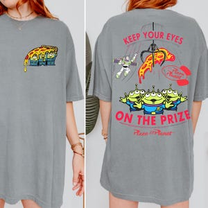 May include: Gray t-shirt featuring a cartoon pizza slice with three green aliens on the front. The back of the shirt has the text "KEEP YOUR EYES ON THE PRIZE" with Pizza Planet graphics and Buzz Lightyear.