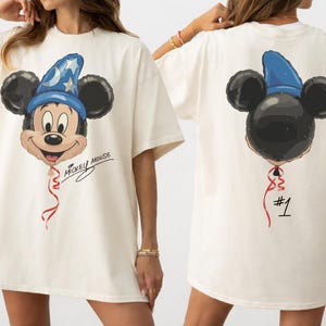 Pode incluir: Uma t-shirt creme com um gráfico de um balão do Mickey Mouse usando um chapéu de feiticeiro azul na frente com o texto Mickey Mouse. As costas exibem o mesmo gráfico do balão visto de trás com o número #1 impresso abaixo.