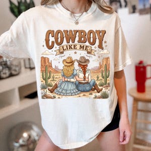Puede incluir: Una camiseta de manga corta color hueso con un estampado gr&aacute;fico de dos personas con sombreros de vaquero sentadas en un paisaje des&eacute;rtico con cactus y mesetas. El texto COWBOY LIKE ME est&aacute; impreso en marr&oacute;n sobre la ilustraci&oacute;n con estrellas.