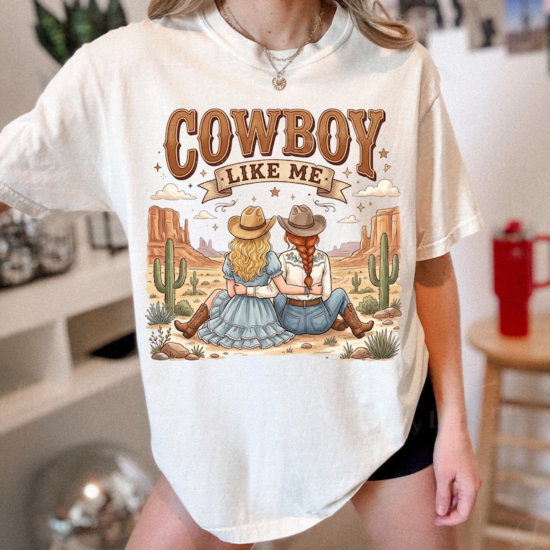 Puede incluir: Una camiseta de manga corta color hueso con un estampado gr&aacute;fico de dos personas con sombreros de vaquero sentadas en un paisaje des&eacute;rtico con cactus y mesetas. El texto COWBOY LIKE ME est&aacute; impreso en marr&oacute;n sobre la ilustraci&oacute;n con estrellas.