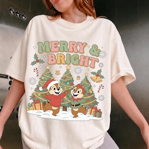 May include: A cream-coloured short-sleeve t-shirt featuring a graphic print with the text MERRY & BRIGHT. The design includes two chipmunks in holiday attire, decorated Christmas trees, snowflakes, holly, candy canes, and gift boxes.