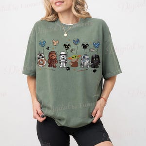 Puede incluir: Camiseta verde salvia con un gr&aacute;fico de personajes de Star Wars sosteniendo globos. Los personajes incluyen a BB-8, Chewbacca, un Stormtrooper, Grogu, R2-D2 y Darth Vader. Los globos tienen dise&ntilde;os de planetas y cabezas de Mickey Mouse.