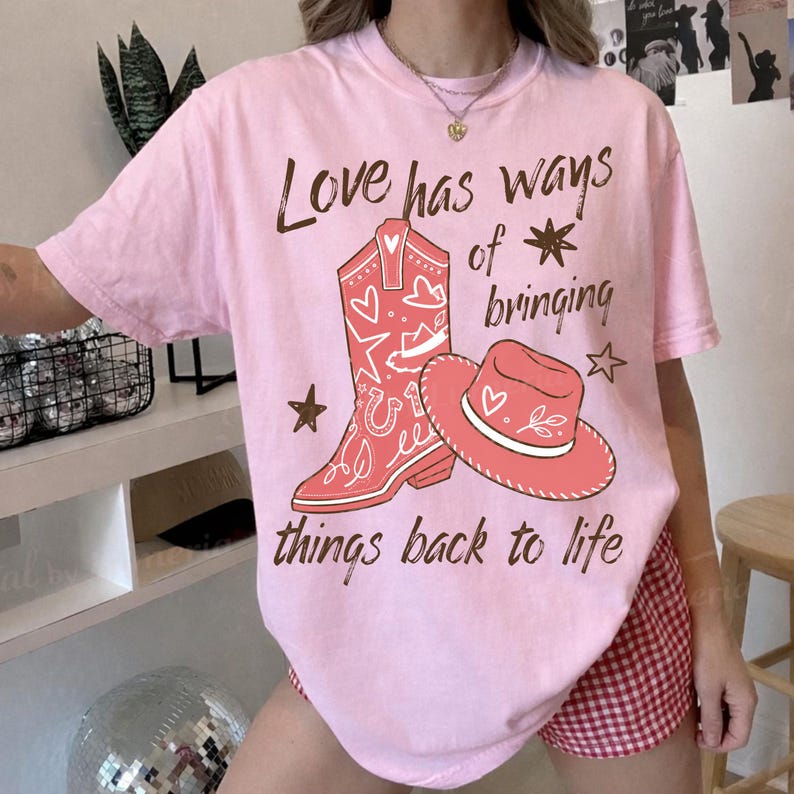 May include: A light pink short-sleeve t-shirt featuring a graphic of a red cowboy boot and a red cowboy hat. The text "Love has ways of bringing things back to life" is printed in brown script around the illustrations, accented by small stars.