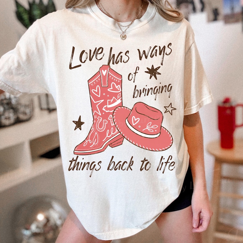 May include: A cream-colored short-sleeve t-shirt featuring a graphic of a red cowboy boot and a red cowboy hat. The text reads, Love has ways of bringing things back to life, accompanied by small star and heart illustrations in a dark brown color.
