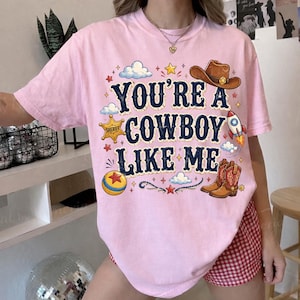 Puede incluir: Una camiseta de manga corta rosa claro con el texto azul oscuro YOU'RE A COWBOY LIKE ME. El gr&aacute;fico incluye un sombrero de vaquero marr&oacute;n, una insignia de estrella de sheriff amarilla, un cohete, una pelota colorida y botas vaqueras marrones.
