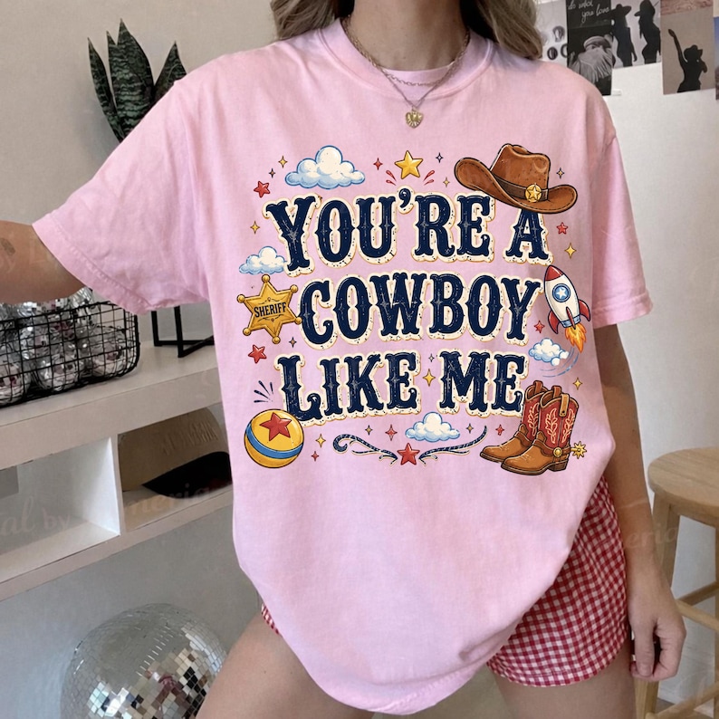 Puede incluir: Una camiseta de manga corta rosa claro con el texto azul oscuro YOU'RE A COWBOY LIKE ME. El gr&aacute;fico incluye un sombrero de vaquero marr&oacute;n, una insignia de estrella de sheriff amarilla, un cohete, una pelota colorida y botas vaqueras marrones.