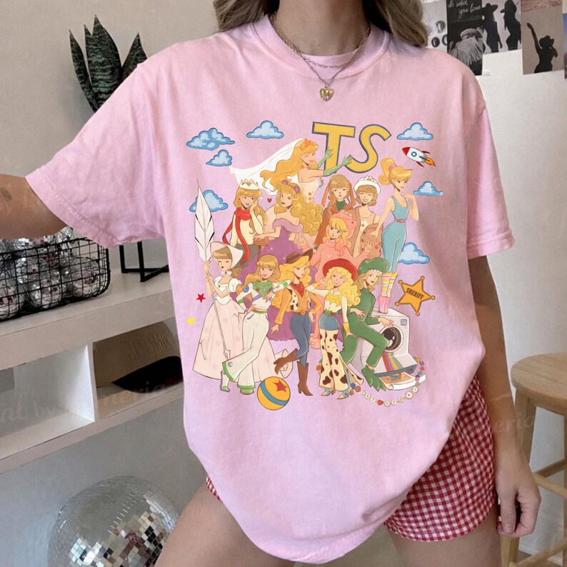 May include: A light pink short-sleeve t-shirt featuring a colorful graphic print of various animated characters in diverse outfits. The design includes the yellow text TS at the top, blue clouds, a small rocket, and a yellow star badge on the front.