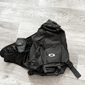 May include: A black Oakley sling backpack featuring a textured fabric construction, a prominent white Oakley logo on the front, multiple zippered compartments, adjustable straps with plastic buckles, and integrated D-rings for additional attachment points.