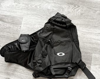 May include: A black Oakley sling backpack featuring a textured fabric construction, a prominent white Oakley logo on the front, multiple zippered compartments, adjustable straps with plastic buckles, and integrated D-rings for additional attachment points.