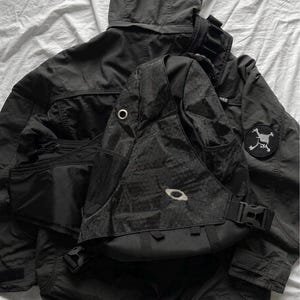 Könnte beinhalten: Eine schwarze Kapuzenjacke mit einem runden Aufnäher am linken Ärmel und ein schwarzer Sling-Rucksack mit strukturiertem Muster, einem einzelnen Schultergurt, seitlichen Netztaschen und einem zentralen gestickten Logo, alles als monochromes Set.