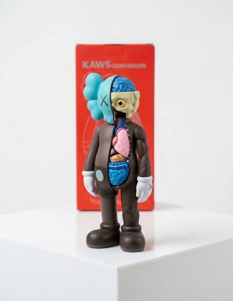 May include: A brown KAWS Companion figure with a partially dissected body, revealing internal organs. The figure has a blue head with X-shaped eyes and a light blue cloud-shaped head. The figure is standing in front of a red box with the text "KAWS COMPANION".
