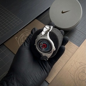 May include: A silver-toned Nike wristwatch featuring a curved, metallic case and integrated link bracelet. The watch face displays a dark circular dial with red accents and a small Nike swoosh logo. It is presented on a black foam cushion.