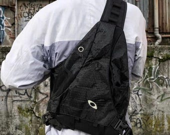 Puede incluir: Una mochila bandolera negra con superficie texturizada, una sola correa ancha para el hombro y un pequeño emblema con el logotipo en blanco. La bolsa cuenta con un ojal metálico, bolsillos laterales de malla y hebillas de plástico negro.